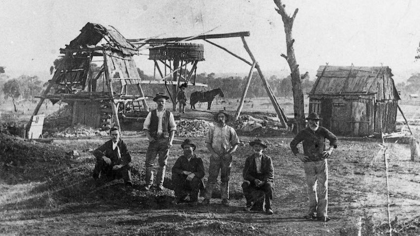 A black-and-white photo of men standing and sitting in a row in front of timber huts, a timber mine shaft, and a horse.