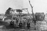 A black-and-white photo of men standing and sitting in a row in front of timber huts, a timber mine shaft, and a horse.