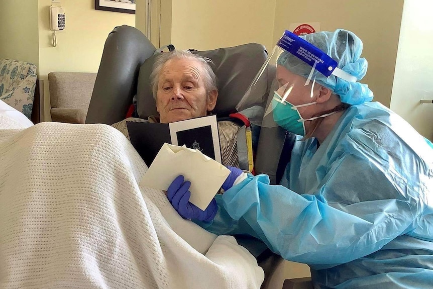 A woman in full PPE sits next to an elderly man who is under a blanket, and reads a card to him.