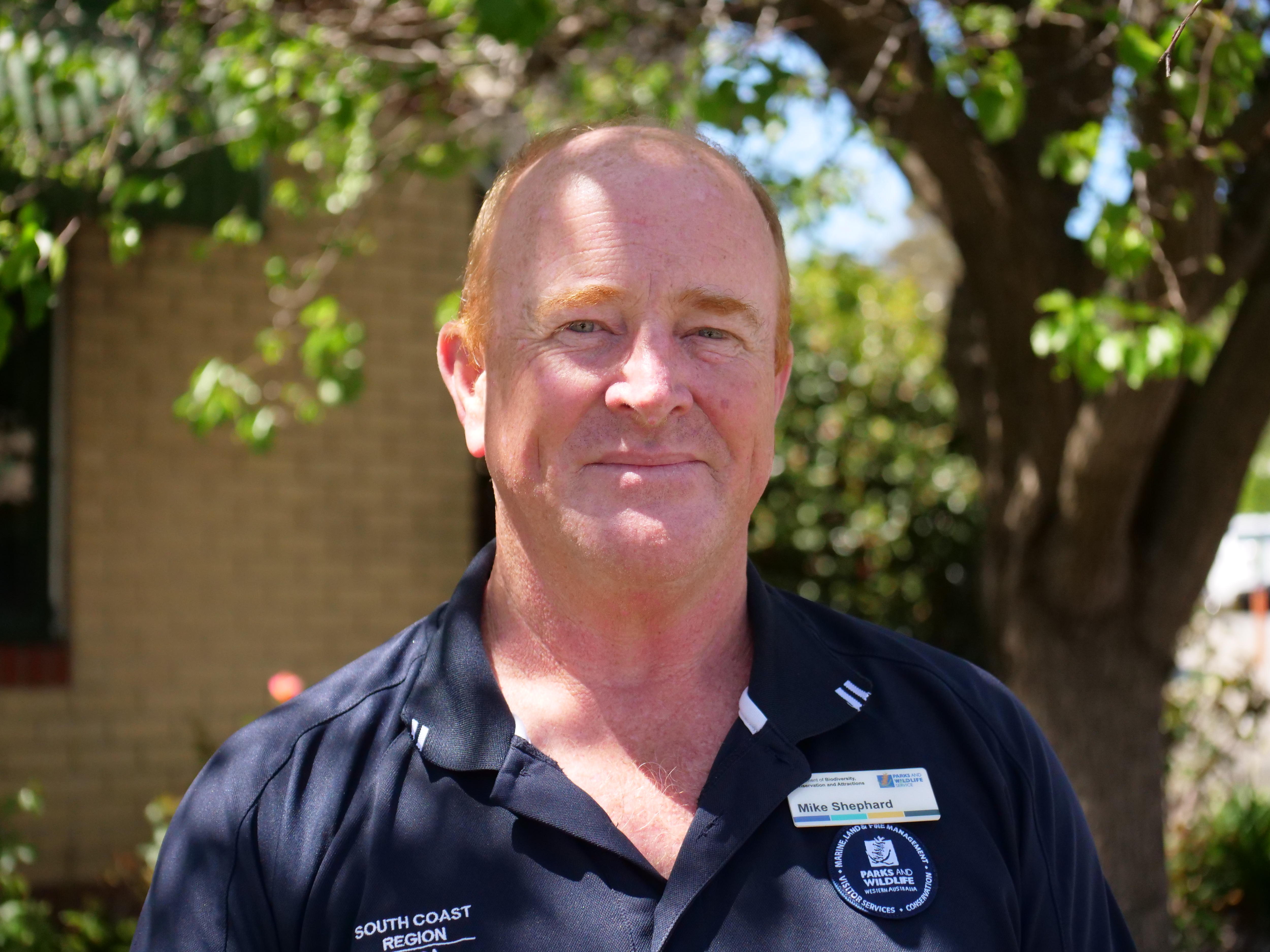 A middle-aged, ginger-haired man wearing a branded polo shirt stands outdoors.