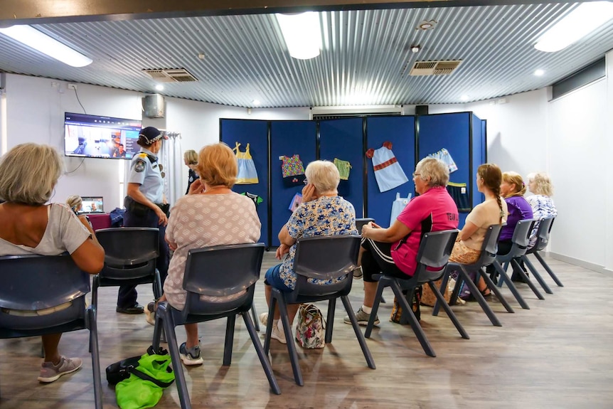 A group of women sit on chairs looking at a screen. Police officers are in the room.