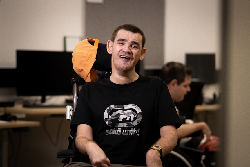 A smiling young man sitting on a wheelchair inside a room, with rows of computers in the background.