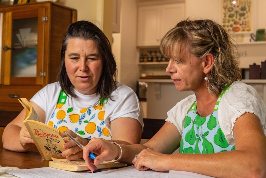 Two woman in colourful aprons sit a a ktichen table looking at an old community cook book.
