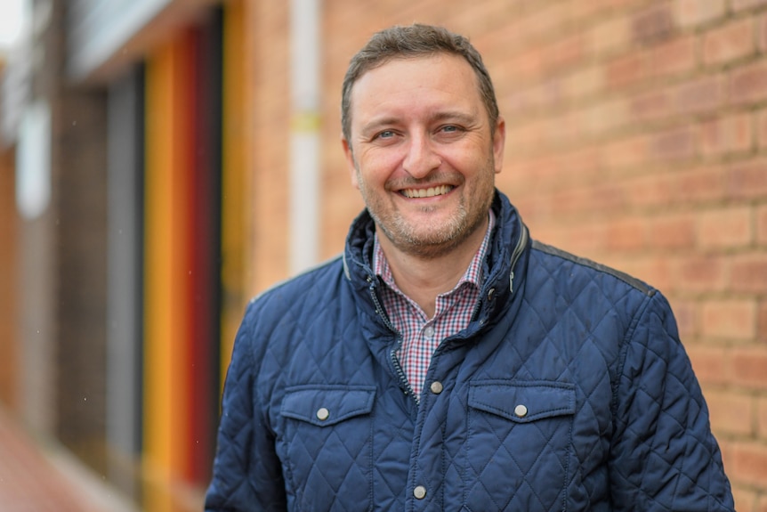 Man stands in front of brick wall smiling while wearing a blue puffer jacket