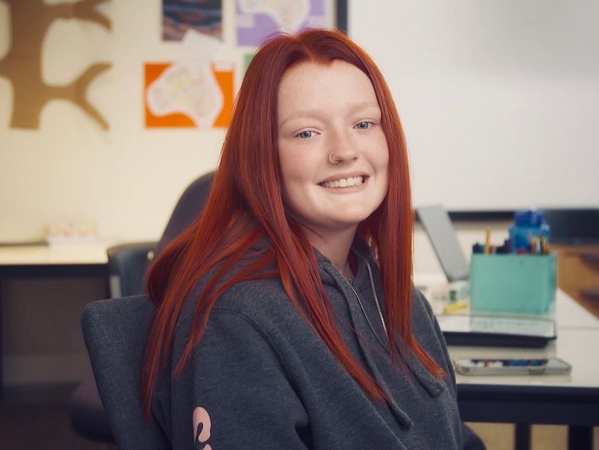 A red-haired student smiles at the camera in a classroom environment