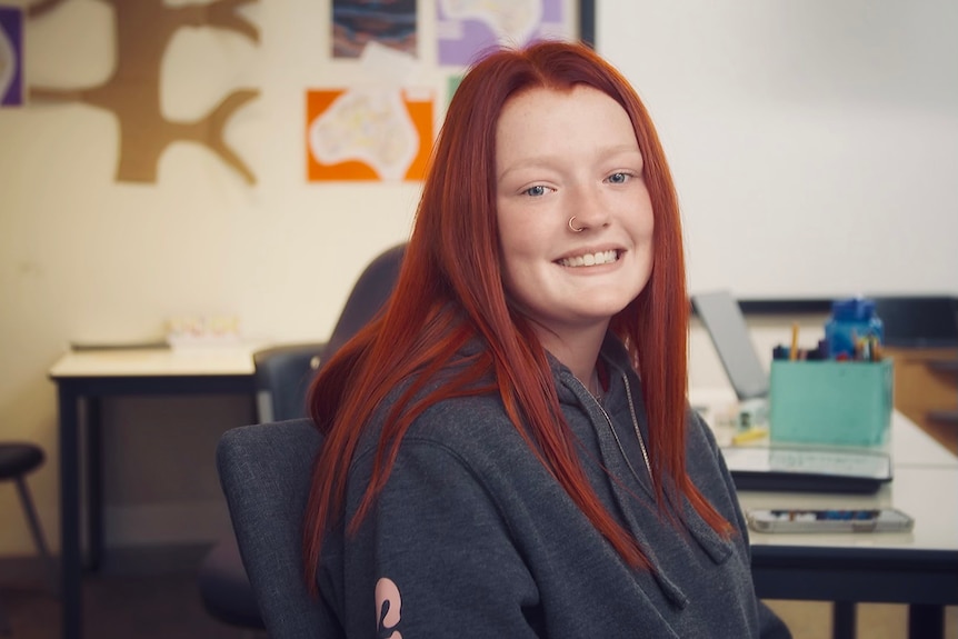 A red-haired student smiles at the camera in a classroom environment