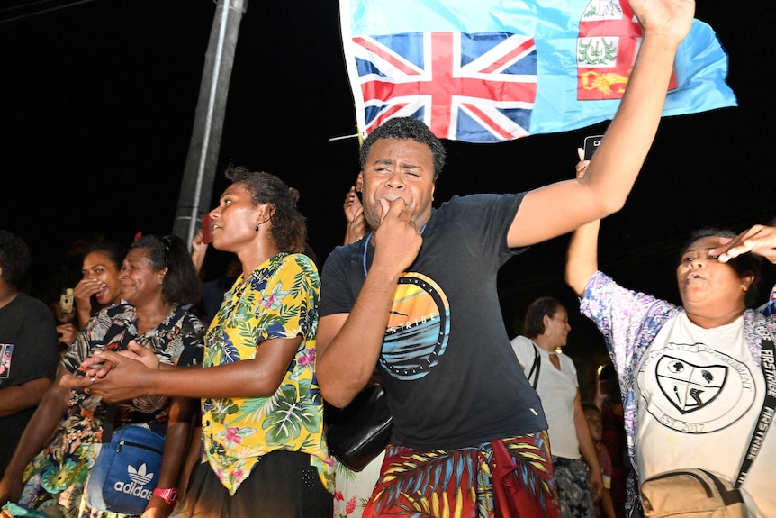 Man whistles and people dance and celebrate in front of a sky blue Fijian flag