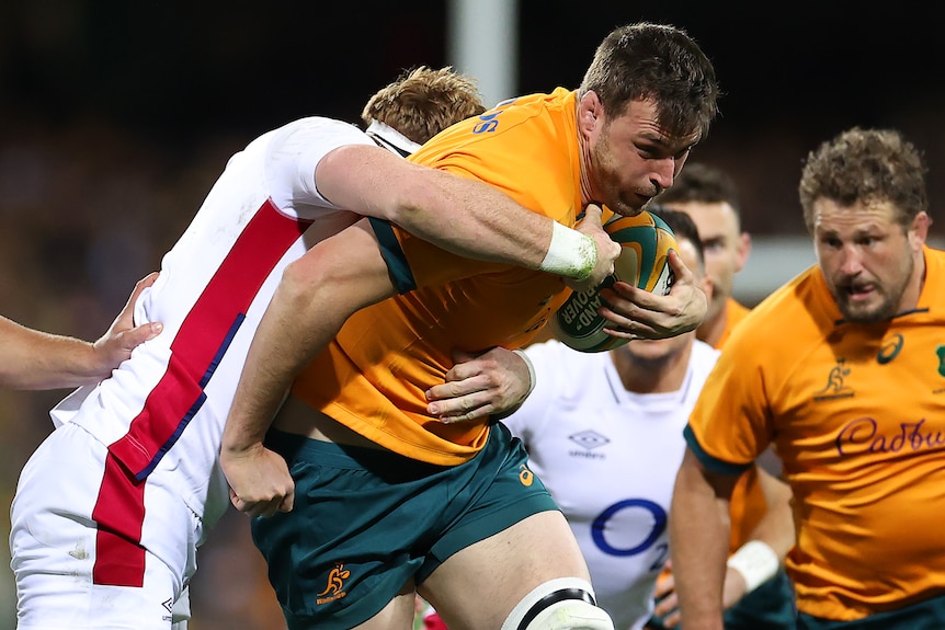 A Wallabies forward holds the ball with his left hand as he is tackled by an English opponent.