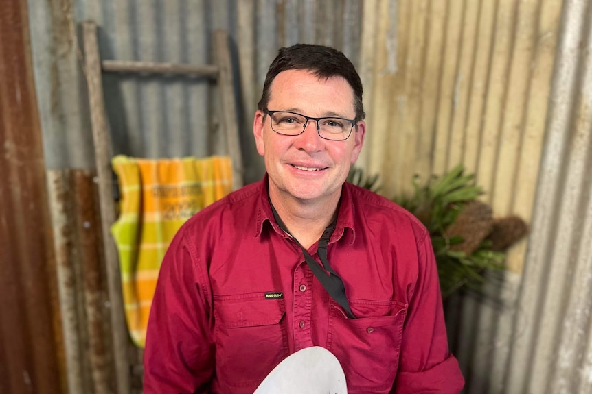 A man stands in a tin shed in a red shirt smiling with glasses on.