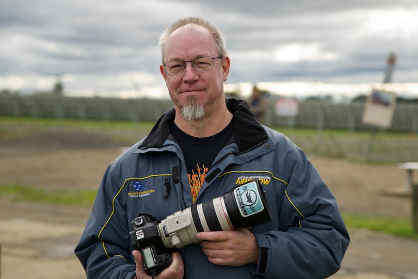 a man with a beard and glasses who is smiling. he is holding a camera.