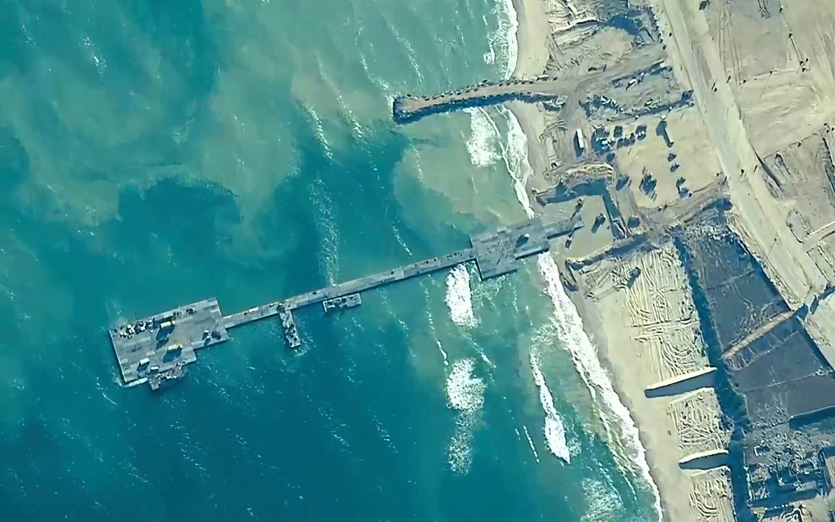 A view from high up shows a grey metal pier extending into the ocean from a narrow, sandy beach.