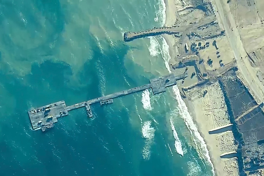 A view from high up shows a grey metal pier extending into the ocean from a narrow, sandy beach.