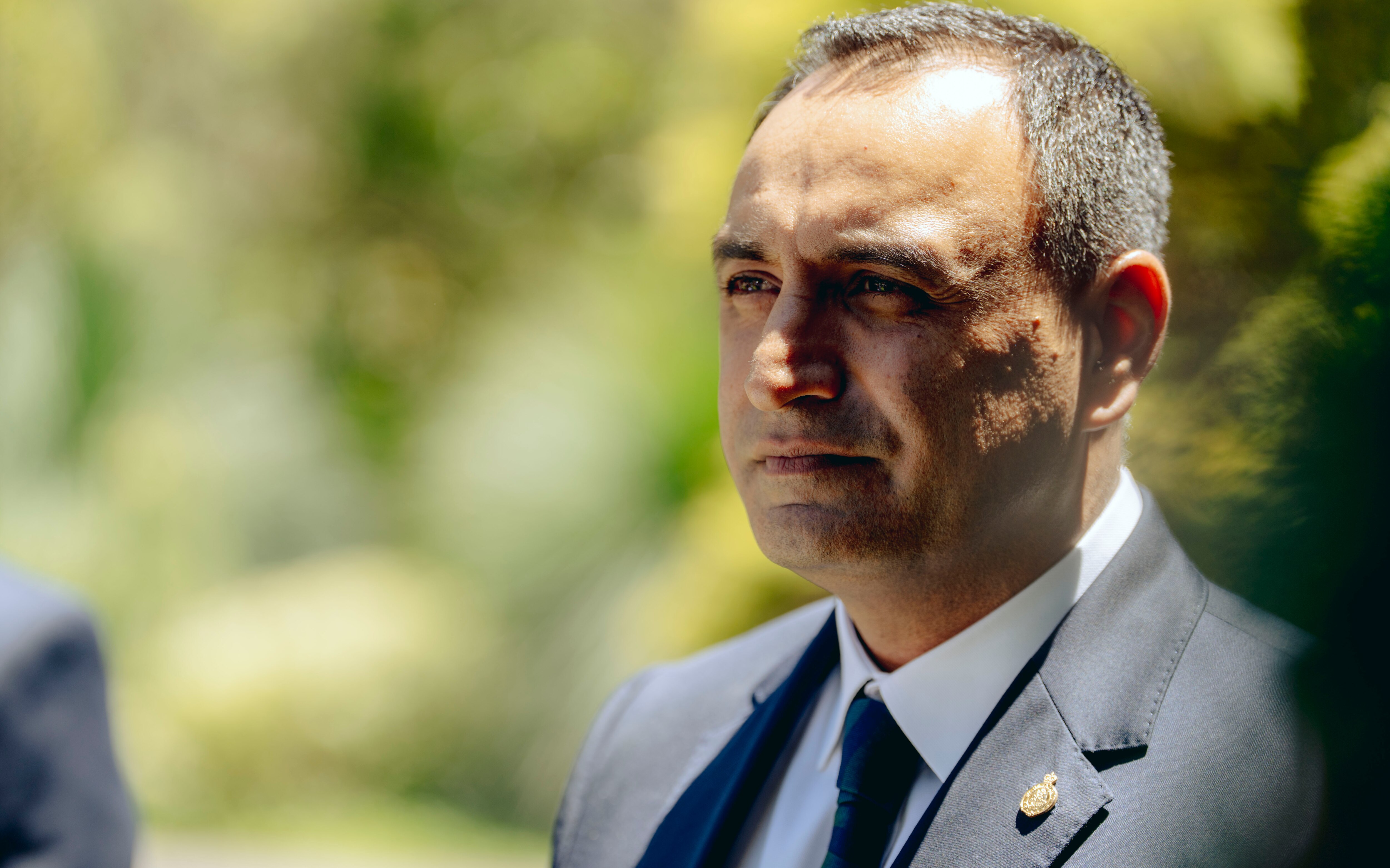 Un hombre mira pensativamente a lo lejos frente a unos arbustos verdes.