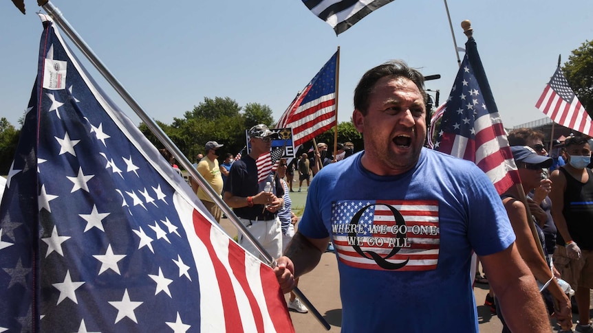 A person wearing a t-shirt supportive of QAnon holds a US flag and screams amid a crowd of demonstrators.