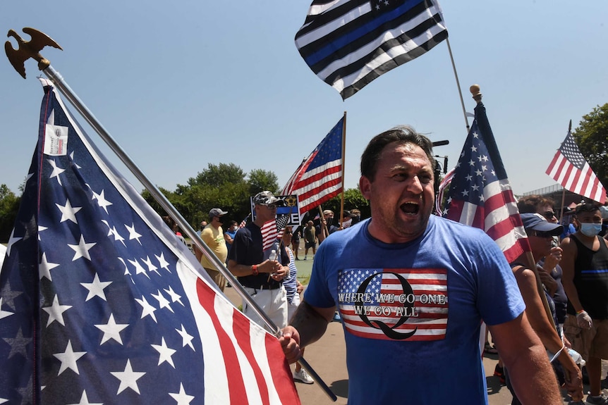 A person wearing a t-shirt supportive of QAnon holds a US flag and screams amid a crowd of demonstrators.