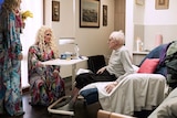 The drag queens talking to a resident in his room, while he sits on his bed.