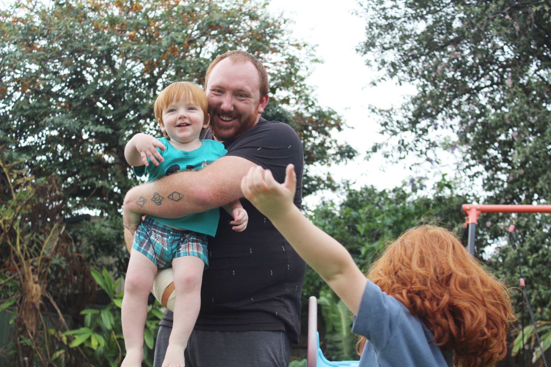 A man smiling and holding his son while his daughter plays in the foreground 