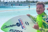 A man wearing a bright yellow and green wetsuit does the hang ten sign with his left hand  in front of a wave pool.