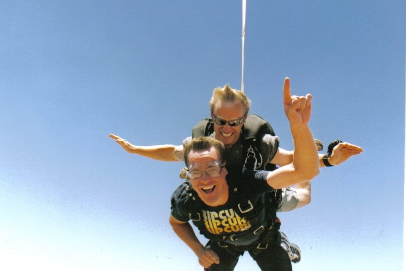 man sky dives in mid air with hand raised with instructor on back