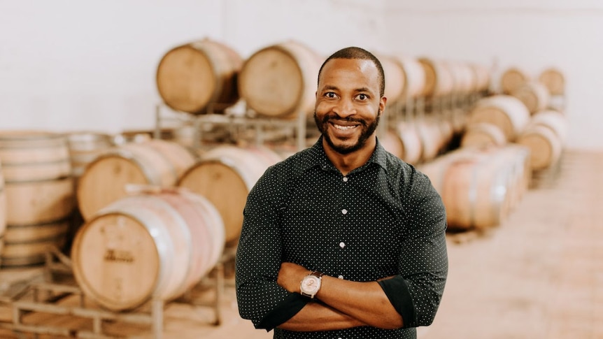 Tinashe Nyamudoka with short black hair and facial hair, stands with arms crossed in front of wine barrels, smiling widely.