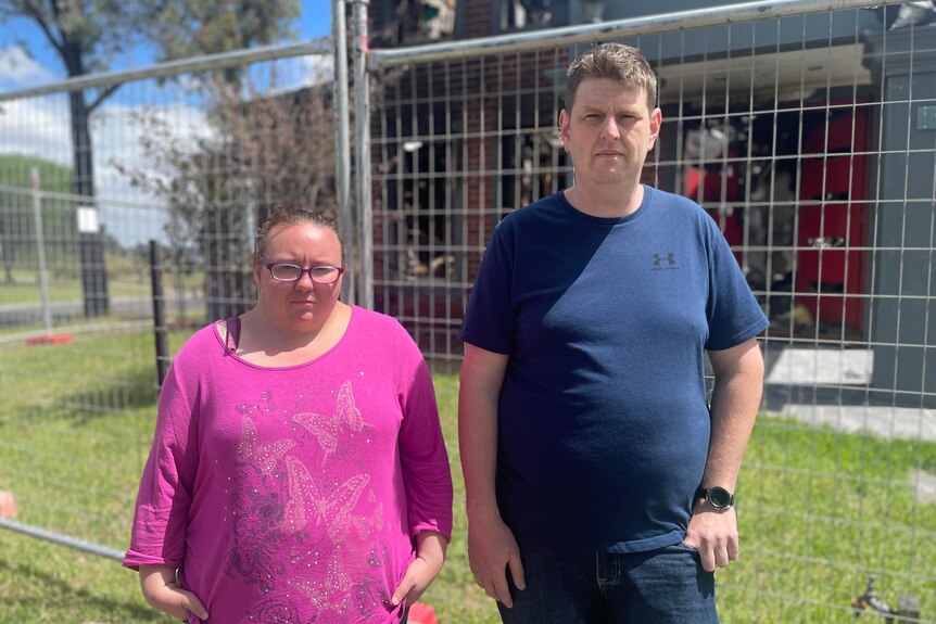 A man and a woman stand in front of a house behind a construction fence. 