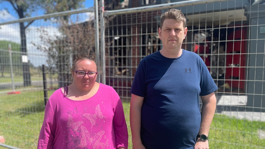 A man and a woman stand in front of a house behind a construction fence. 