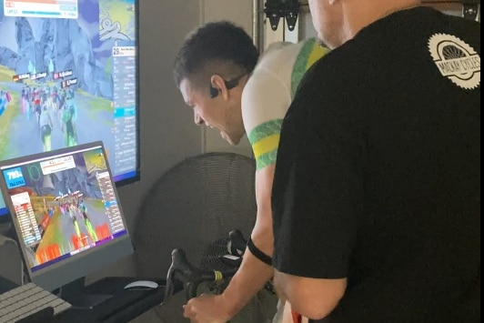 A man on a stationary bike in front of a laptop and TV screen with a man in a black shirt standing next to him