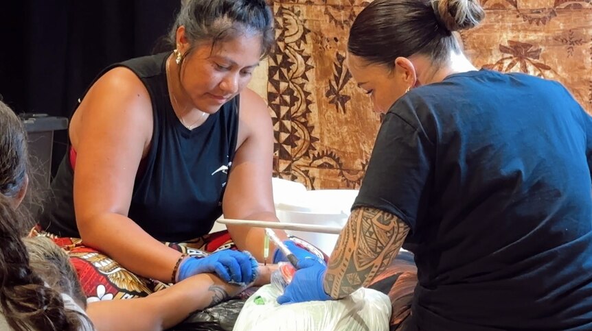 Two women can be seen wearing blue gloves giving a hand a traditional tualima tatau with the 'au. 