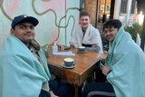 Three young men wrapped in blankets sit at a cafe table and drink coffee.