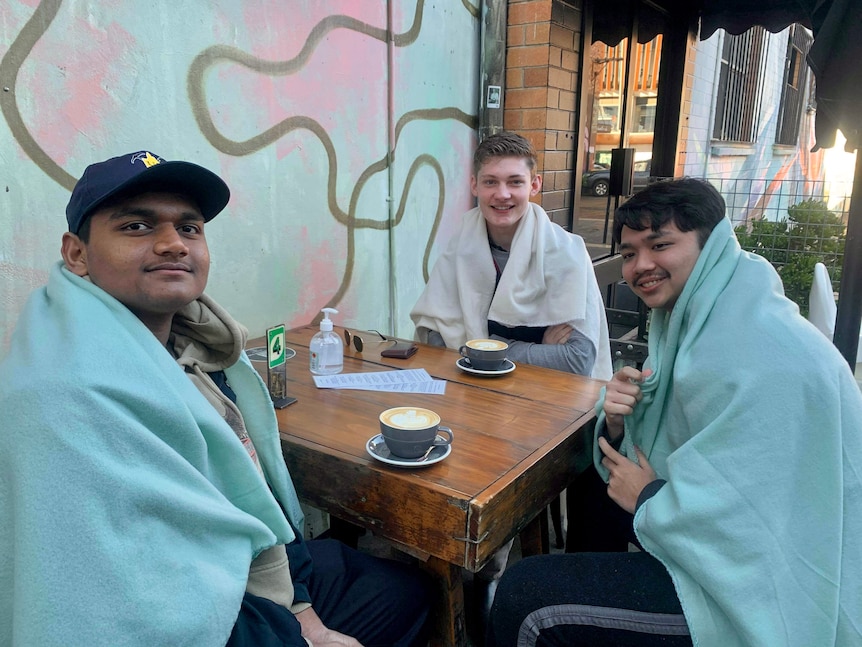 Three young men wrapped in blankets sit at a cafe table and drink coffee.