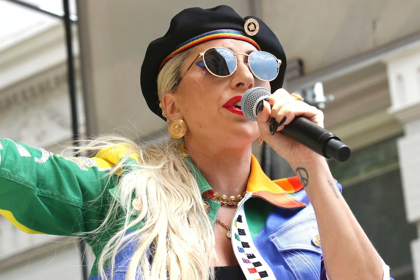 Lady gaga wearing a rainbow jacket and har holds a microphone to her mouth