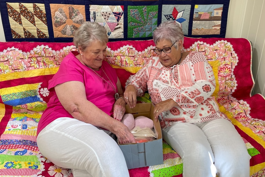 Two women sit on a brightly coloured couch with some knitted breasts, with a quilt hanging behind.