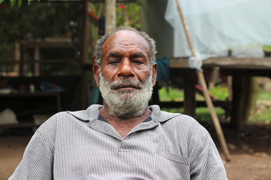 A man sitting outdoors stares at the camera, a neutral expression on his face.