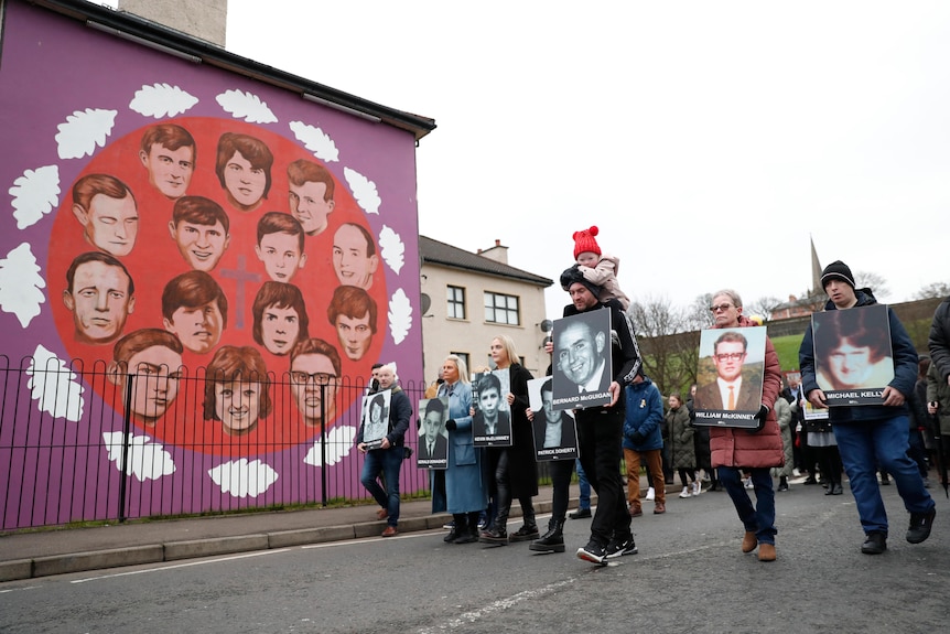 Oameni care dețin fotografii cu victime trec pe lângă o pictură murală care înfățișează victimele Duminica Sângeroasă