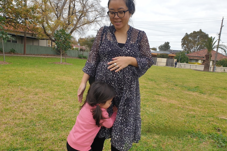 Pregnant woman stands in a park as her preschool daughter cuddles her legs