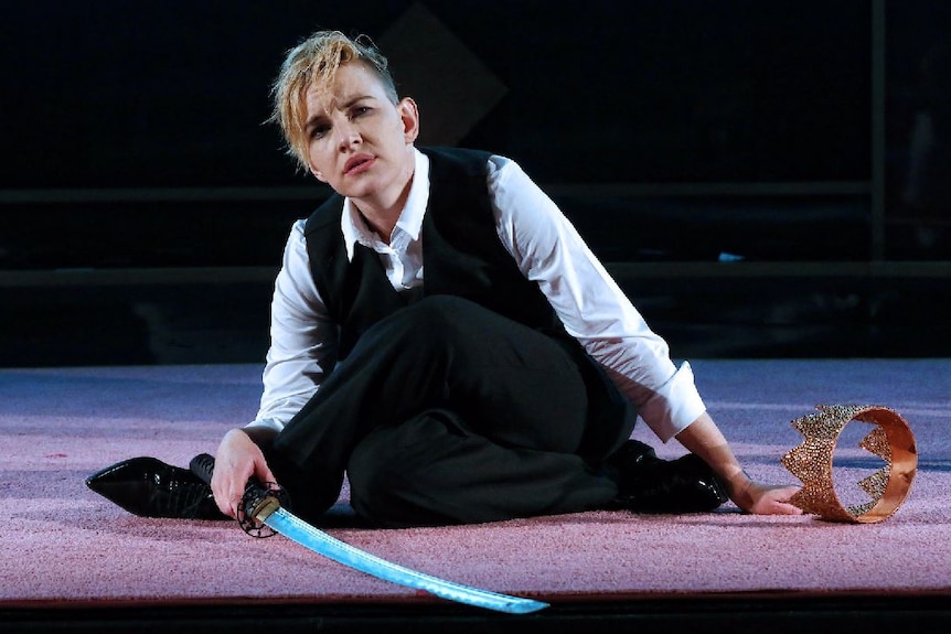 A woman with blonde hair sits on a stage, wearing a white shirt, black vest and black pants.