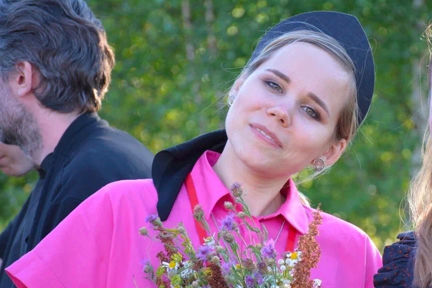 A young blonde woman in a black headband, a neon pink dress, clutching a bunch of flowers to her chest 