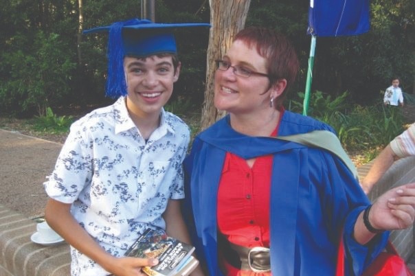 boy looks at camera smiling and wearing a royal blue graduation hat. His mother sits next to him wearing a graduation robe