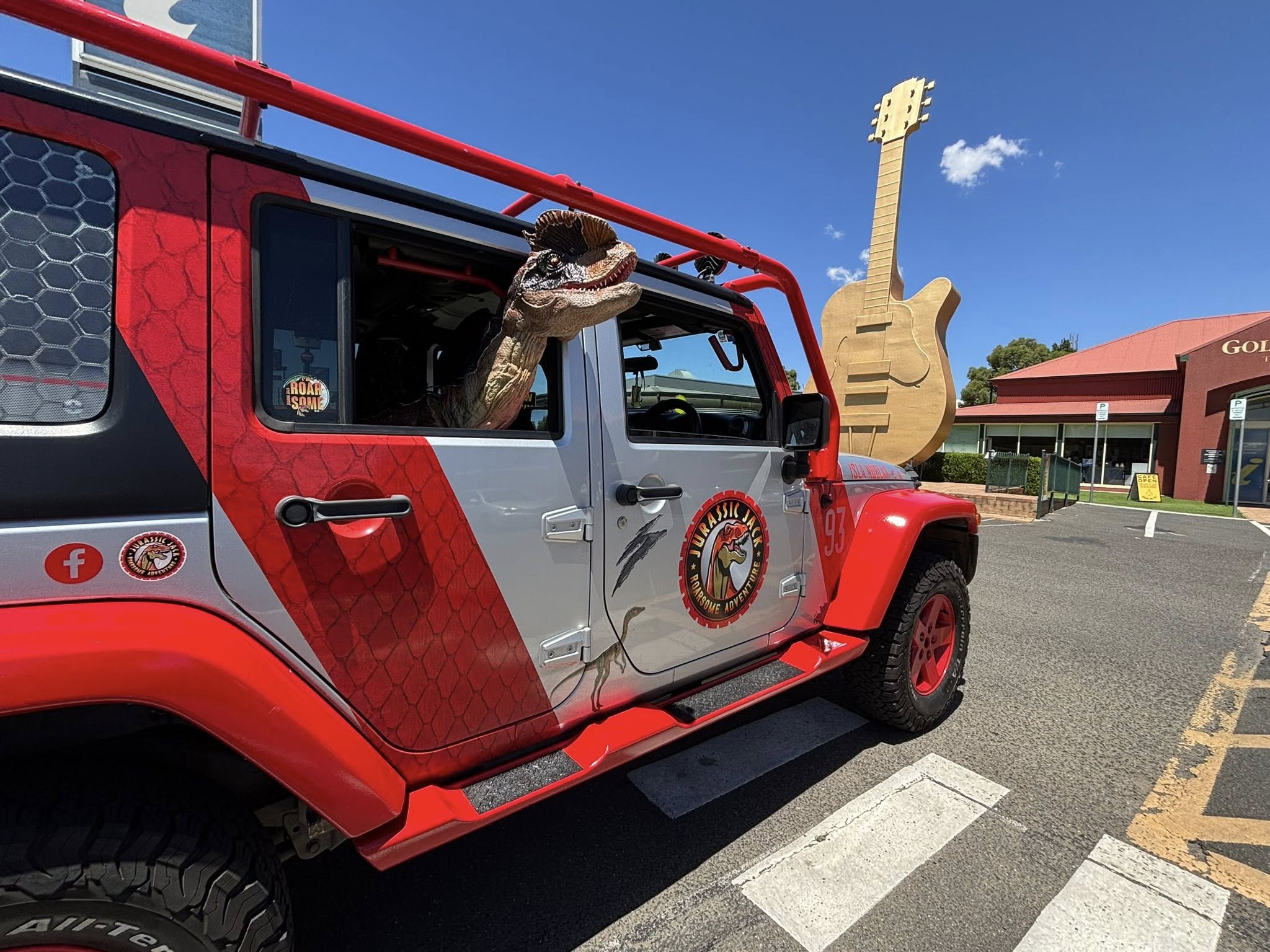 Un modelo de dinosaurio asoma la cabeza por la ventana trasera de un vehículo de Jurassic Park frente a una escultura de guitarra gigante