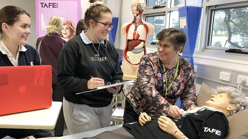 Tracey Newcombe demonstrates a technique on an elderly mannequin in a bed while two students watch on.