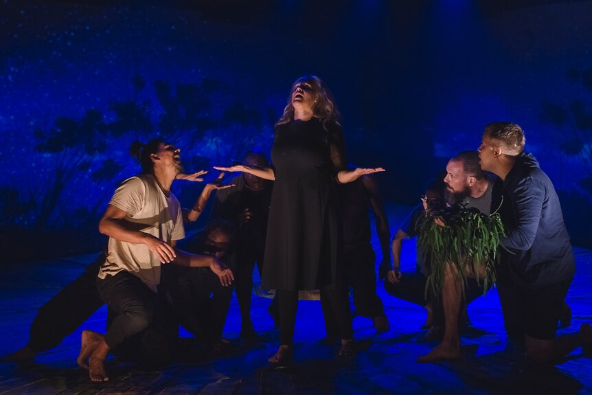 Darkened stage with night skyline with trees projected in background, and group of 8 crouched around woman standing arms out.