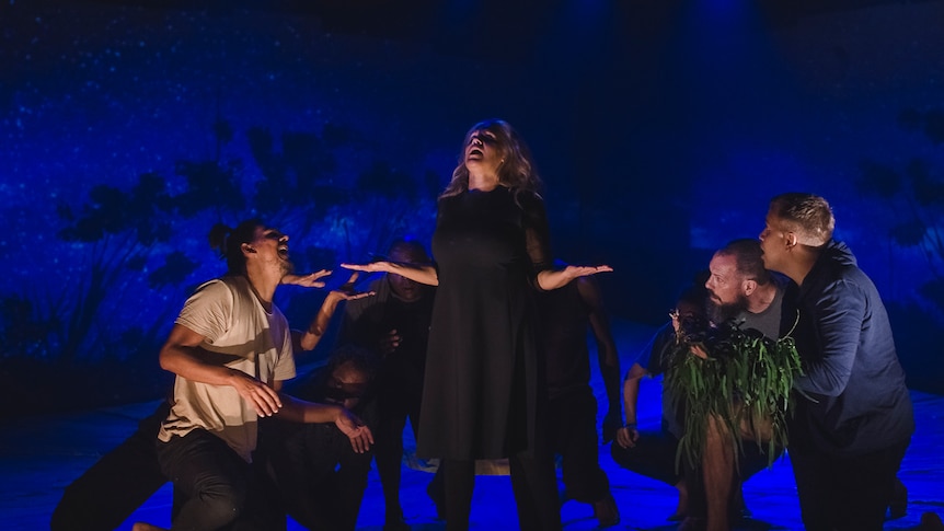 Darkened stage with night skyline with trees projected in background, and group of 8 crouched around woman standing arms out.