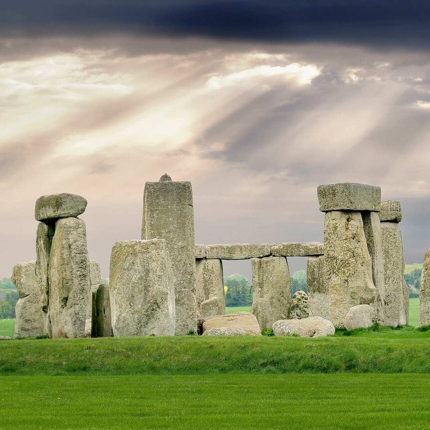 Tall standing stones at Stonehenge in the UK, photographed against a dramatic backdrop of sun shining through dark clouds.
