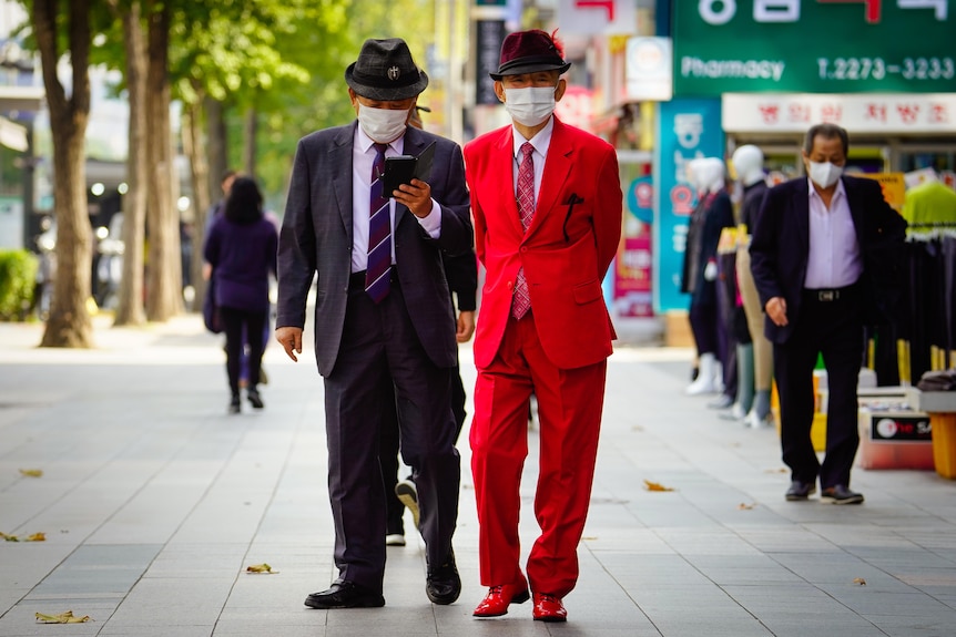 Two older Korean men, one wearing a bright red suit and hat, and the other in a dark-colored suit and hat