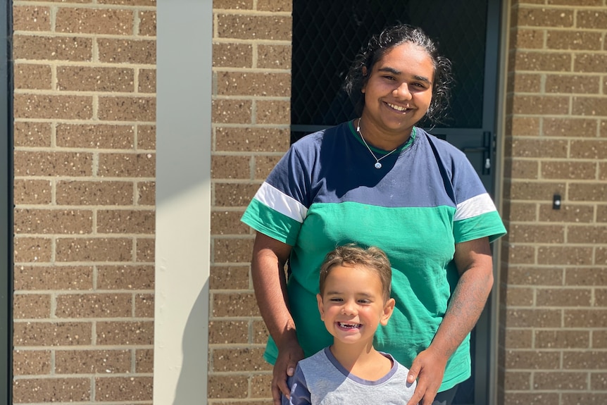 Mother standing out the front of her new home smiling with her four year old son. 