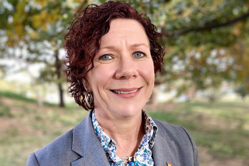 A woman with short curly hair, wearing a suit jacket with a small rainbow pin, smiles at the camera.