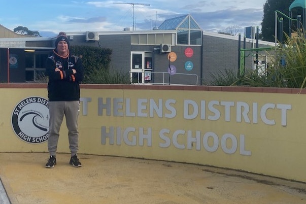 A man in a beanie stands with folded arms in front of St Helens District High School 