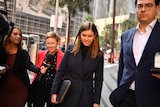 Brittany Higgins walks through a crowd of journalists carrying a folder, wearing a black coat.