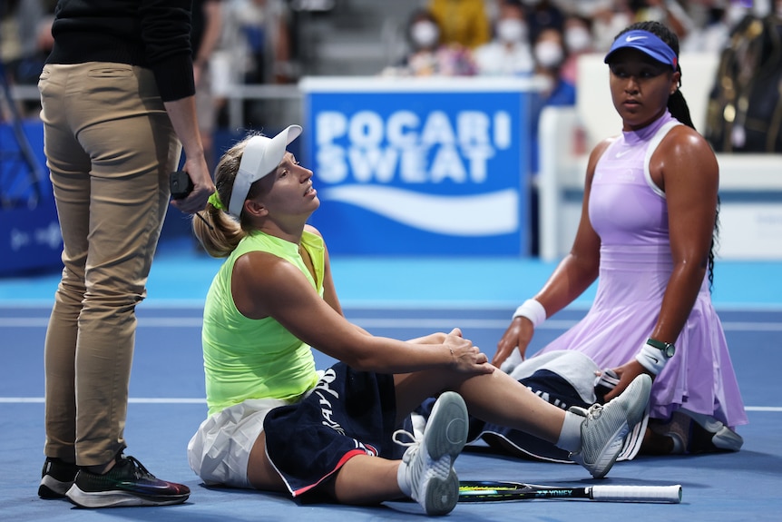 Naomi Osaka kneels next to Daria Saville as she clutches her injured knee on a tennis court