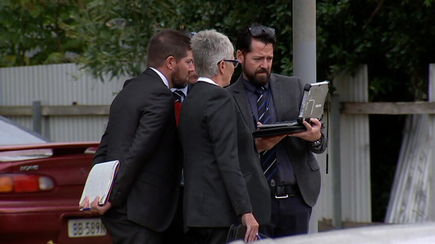 A group of detectives wearing suits huddle around an ipad at the scene of an assault